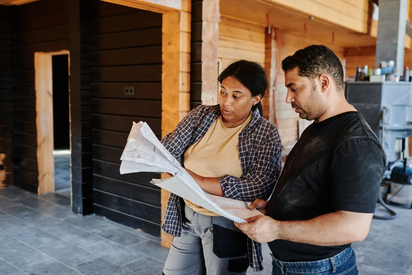 Two construction workers review blueprints inside a wooden structure, discussing plans
