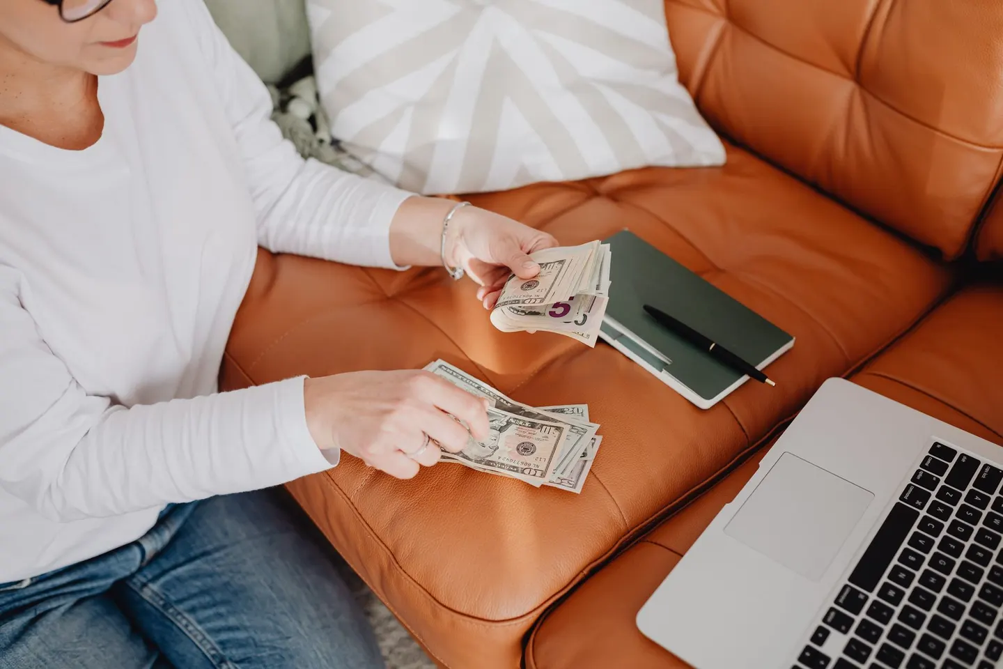 Woman counting cash on a sofa beside a laptop and notebook