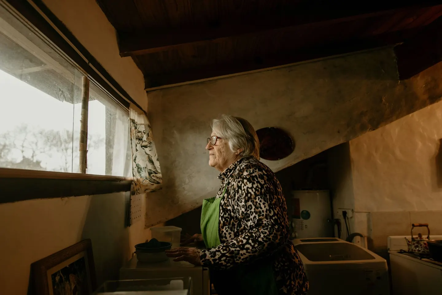 An elderly woman wearing glasses and a leopard print jacket looks out a window indoors